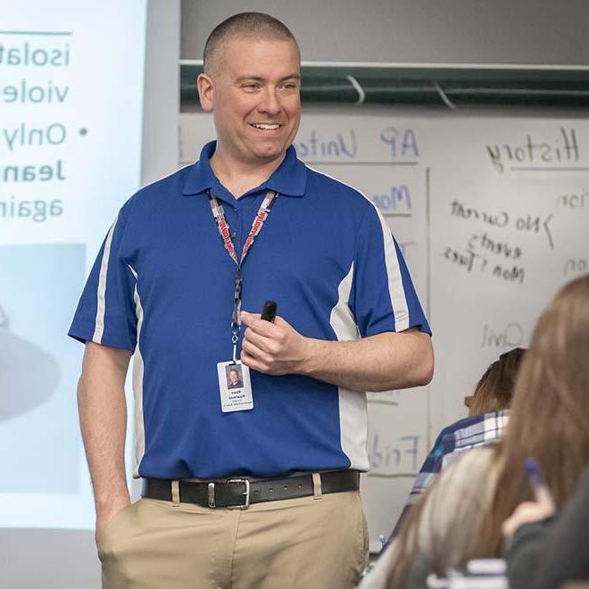 High school teacher smiling in front of classroom with presentation behind him