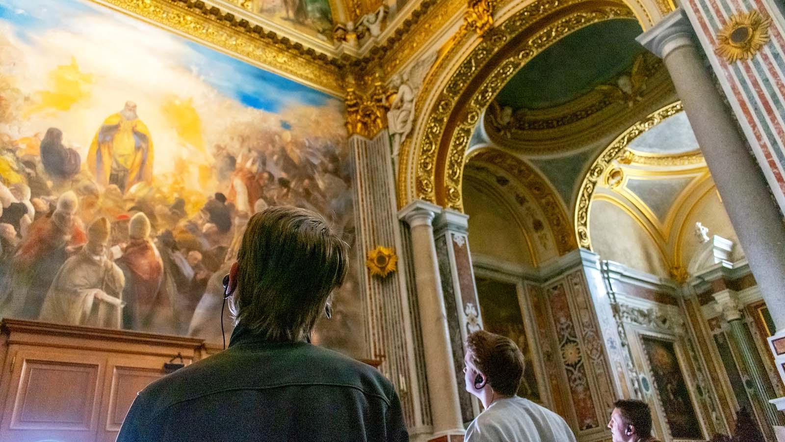 Three students studying abroad in Rome listening to commentary on headsets while admiring a classical painting in a museum