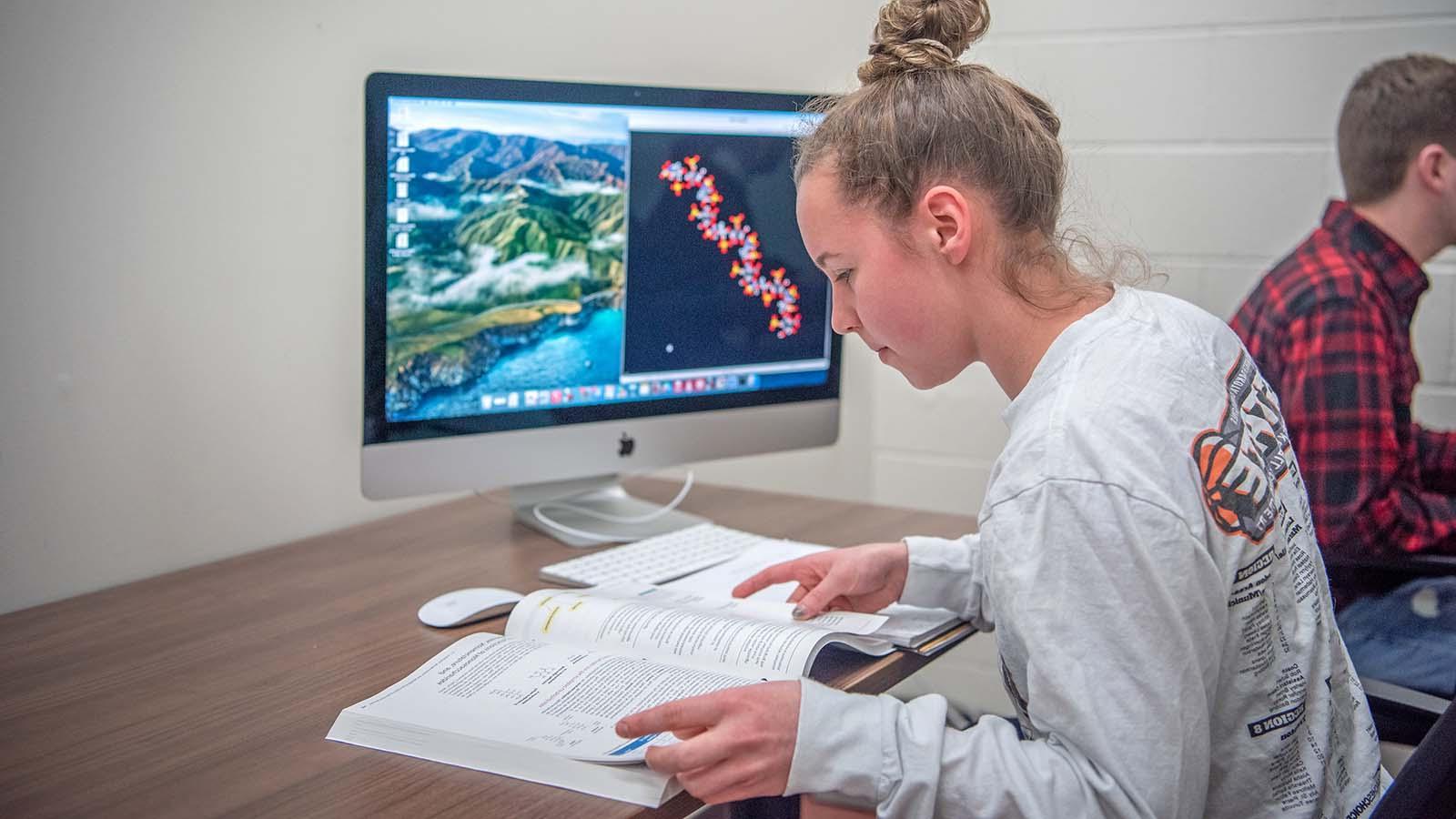Female student looking at textbook with computer in front of her with DNA strand on the screen.